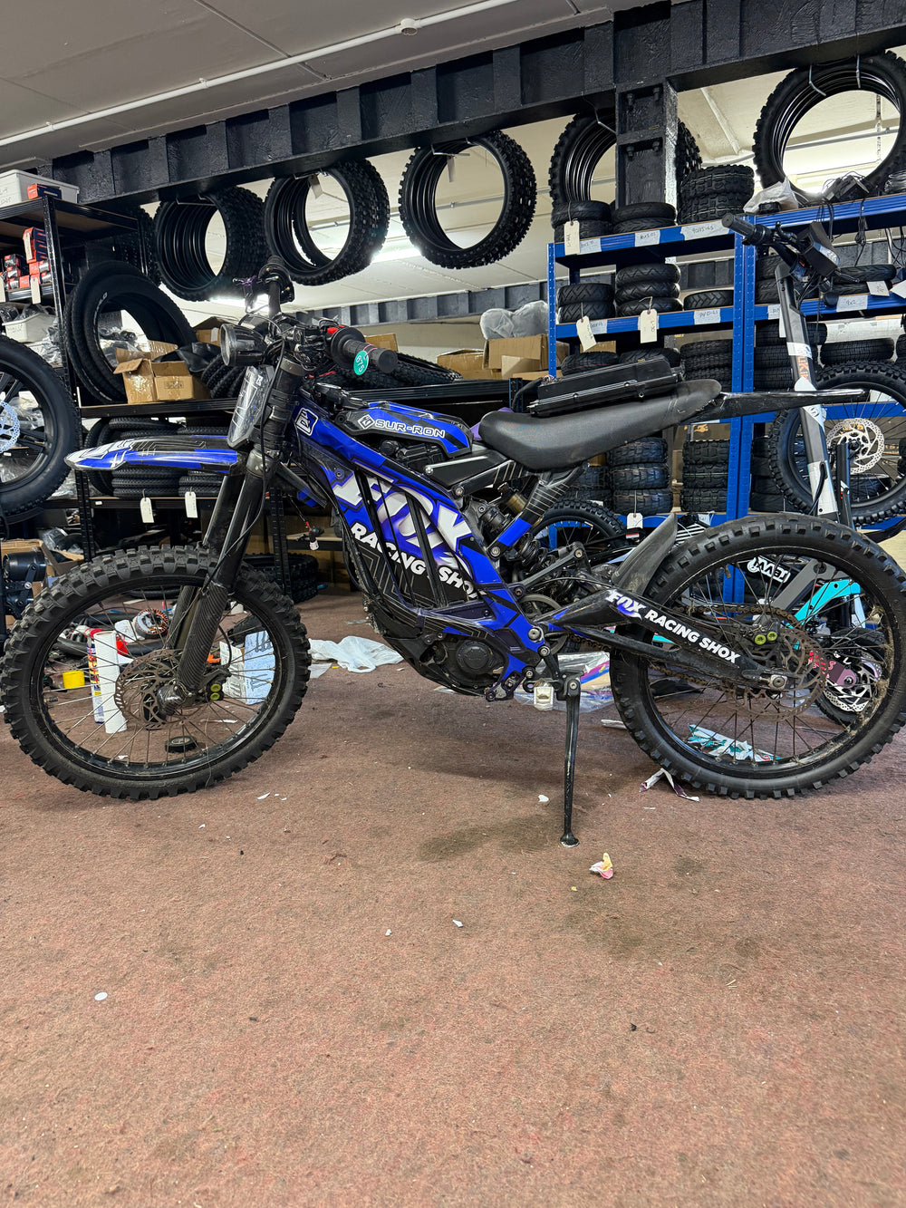 Blue Sur-Ron electric bike beside tyre and parts shelving in the Yocks workshop